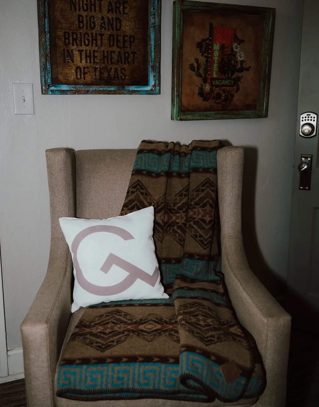 A beige upholstered armchair with a Southwestern-patterned brown and turquoise blanket draped over it. A white accent pillow with a large stylized “G” logo rests on the seat. Behind the chair, two rus